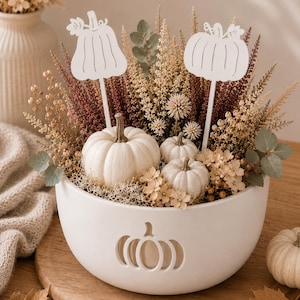 Puede incluir: Un cuenco de cerámica blanco con un diseño de calabaza recortado, lleno de flores secas, hierbas y tres pequeñas calabazas blancas. Dos marcadores blancos con forma de calabaza en palitos están insertados en el arreglo, que descansa sobre una tabla.