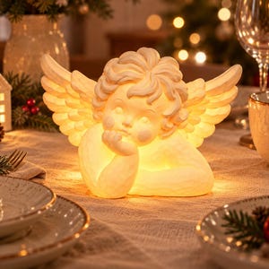 May include: A cream-colored, illuminated angel figurine featuring a cherubic face with curly hair, large feathered wings, and a hand resting under the chin. The sculpture emits a warm, soft glow from within, highlighting its textured surface.