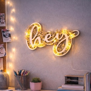May include: A wooden wall decoration spelling "hey" with warm white LED lights. The word is surrounded by delicate floral accents. The piece is mounted on a light grey wall, with a desk and accessories in the foreground.