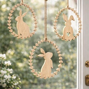 May include: Three wooden Easter bunny wreaths hanging in front of a window. Each wreath features a bunny silhouette in the centre, surrounded by a heart-shaped border. The wreaths are suspended by twine.