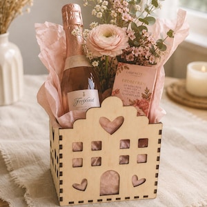 Könnte beinhalten: Eine holzförmige Geschenkbox in Hausform mit einer Flasche Freixenet Sekt, rosa Seidenpapier, einem rosa Blumenarrangement und einer Schokoladentafel. Die Box hat herzförmige Ausschnitte und quadratische Fenster.