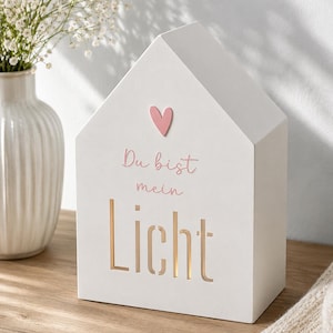 May include: A white house-shaped wooden block with a pink heart icon and the text "Du bist mein Licht" printed on the front. The word "Licht" is cut out, allowing light to shine through the opening, creating a warm glow from within the structure.
