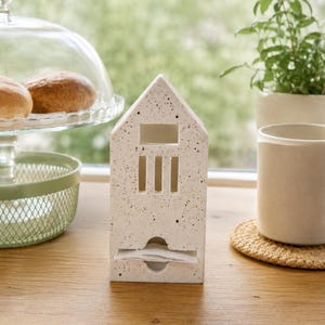 May include: A house-shaped tissue dispenser made of off-white material with dark brown speckles. The front features a rectangular opening at the top, three vertical slits in the middle, and a horizontal slot at the bottom for dispensing tissues.