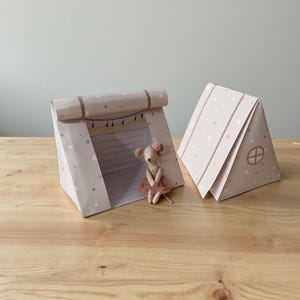 Puede incluir: Dos tiendas de campaña de papel, una abierta y otra cerrada, en color beige claro con un estampado de lunares. La tienda abierta tiene una guirnalda de luces y una pequeña muñeca ratón con un tutú rosa. La tienda cerrada tiene un diseño de ventana pequeña.