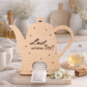 Può includere: Porta bustine di tè in legno a forma di teiera, con il testo tedesco "Lust auf einen Tee?" e punti decorativi. Il supporto poggia su una base di legno, con bustine di tè visibili. Una tazza di tè e fiori sono nelle vicinanze.