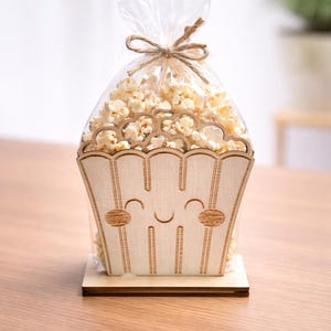 Könnte beinhalten: Ein hölzerner Popcornbehälter mit einem lächelnden Gesicht, gefüllt mit Popcorn und in einer transparenten Plastiktüte mit Bindfaden verschlossen. Der Behälter steht auf einem Holzsockel, ideal für eine Party oder einen Filmabend.