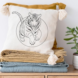 Könnte beinhalten: Ein weißes Zierkissen mit einer schwarzen Tigerillustration in einem Kreis. Es liegt auf einem Stapel gefalteter Decken in Blau- und Beigetönen. Rechts ist eine kleine Topfpflanze zu sehen. Das Kissen hat Quasten.