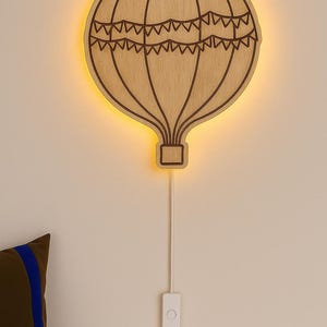 May include: Wooden hot air balloon wall light with a warm glow. The light is made of light brown wood with a dark brown outline and a decorative banner design. It is connected to a white cord and switch.
