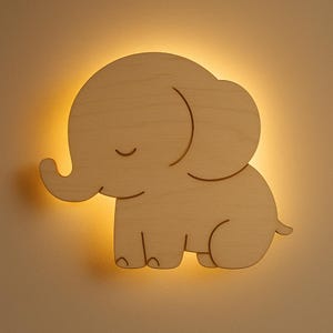 Puede incluir: Una lámpara de pared con forma de elefante de madera, con un brillo amarillo cálido que emana de detrás del diseño recortado. El elefante está hecho de madera de color claro, con un diseño simple y minimalista, perfecto para una habitación infantil.