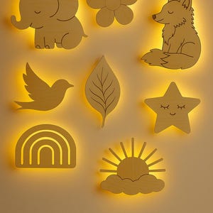 Op de afbeelding: Houten wanddecoratie met een olifant, bloem, vos, vogel, blad, regenboog, ster en zon. Elk stuk is uit hout gesneden en heeft een warme, gele gloed van achteren, waardoor een zachte, uitnodigende sfeer ontstaat.