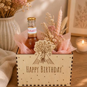 Könnte beinhalten: Eine Holzkiste mit der Aufschrift "HAPPY BIRTHAY" auf der Vorderseite. Die Kiste enthält eine Flasche Lillet, Trockenblumen, eine rosa Kerze und ein dekoratives Holzbouquet. Die Kiste ist mit rosa Seidenpapier gefüllt.