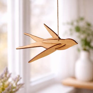 Op de afbeelding: Houten zwaluw vogeldecoratie hangend aan een touwtje. De vogel is gemaakt van lichtgekleurd hout met donkerbruine randen en hangt voor een raam. De vleugels van de vogel zijn uitgespreid, wat de indruk van vlucht geeft. Een decoratief item.