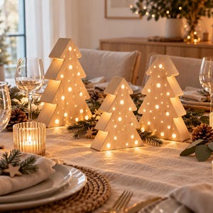 May include: Three light-colored wooden Christmas tree decorations with cut-out circular holes that emit a warm, glowing light. The trees are arranged on a dining table among pine branches, pinecones, and place settings with glassware.