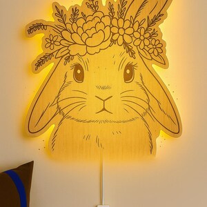 Puede incluir: Luz de pared de madera con forma de conejito y corona floral. La luz es de color amarillo cálido y los rasgos del conejo están grabados en la madera. Tiene un cable de alimentación con un interruptor de encendido/apagado. Decoración infantil.