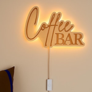 Könnte beinhalten: Holzschild "Coffee Bar" mit warmer LED-Hintergrundbeleuchtung. Das Schild ist an einer Wand befestigt und hat ein Netzkabel mit Ein-/Ausschalter. Das Schild besteht aus hellem Holz und zeigt die Worte "Coffee Bar" in einer stilisierten Schriftart.
