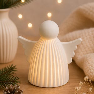 May include: A white, matte-finish angel figurine with a spherical head and a pleated, bell-shaped body. The angel features outstretched wings and emits a warm, soft internal glow from within its structure, creating a gentle light effect.