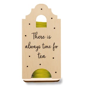 May include: Wooden tea bag holder with the phrase "There is always time for tea" in black script. The holder is light brown with black dots and a curved top. Inside, a stack of yellow tea bags is visible.