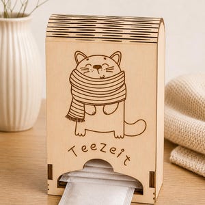 May include: Wooden tea bag dispenser featuring a laser-cut cat design wearing a scarf. The word "Teezeit" is written below the cat. The dispenser holds tea bags and is on a wooden surface.