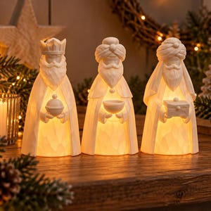 May include: Three white, illuminated figurines representing the Three Wise Men. Each figure features a carved, textured surface, a long robe, and a beard. They hold distinct objects: a lidded vessel, a small bowl, and a rectangular box.