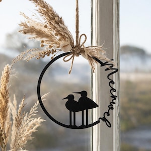May include: A black metal wall decoration with two bird silhouettes inside a circular frame. The frame is decorated with dried flowers and a jute string for hanging. The word "Meerweh" is written in cursive on the frame.
