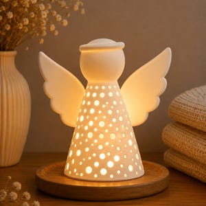 May include: A white ceramic angel figurine with a smooth finish and a halo, featuring a perforated body with circular cutouts that emit a warm internal glow. The angel is positioned on a circular wooden base, showcasing a conical shape with wings.