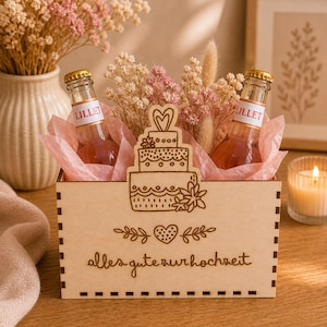 Könnte beinhalten: Eine Holzkiste mit der Gravur "alles gute zur hochzeit" auf der Vorderseite, die zwei Flaschen Lillet enthält. Die Kiste ist mit einem lasergeschnittenen Kuchen- und Herzdesign verziert und mit rosa Seidenpapier und Trockenblumen gefüllt. Im Hintergrund eine brennende Kerze.