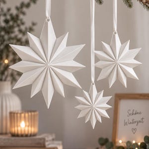 Op de afbeelding: Drie matwitte stervormige ornamenten met meerdere spitse punten, elk opgehangen aan een dun wit lint. De sterren variëren in grootte en hebben een geometrisch, gefacetteerd ontwerp met scherpe randen, hangend aan een tak tegen een neutrale achtergrond.