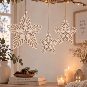 May include: Three white, laser-cut wooden star ornaments with intricate, filigree-style patterns hanging from a natural branch by thin twine strings. Each star features a central five-pointed shape surrounded by layered, curved, petal-like cutouts.