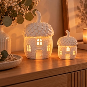 May include: Two white ceramic acorn-shaped lanterns with textured caps and smooth bodies. Each lantern features a central arched door and multiple small windows that emit a warm, glowing light from within, creating a cozy, illuminated display.