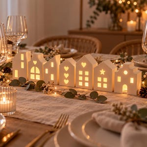 Pode incluir: Uma fila de sete suportes para velas de chá em cerâmica branca em forma de casa dispostos sobre uma mesa. Cada casa apresenta janelas, portas e formas recortadas como corações e estrelas, iluminadas por dentro com um brilho quente e suave.