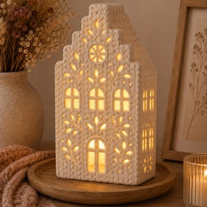 May include: A cream-colored ceramic house-shaped lantern with a textured cable-knit pattern on the exterior. The structure features cut-out windows and a circular attic vent that emit a warm, soft glow from an internal light source.