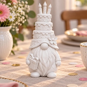 May include: A white gnome figurine featuring a tall, tiered birthday cake hat topped with three candles. The gnome has a long beard, a round nose, and holds a small gift box with a bow in one hand. A single flower is attached to the side of the hat.