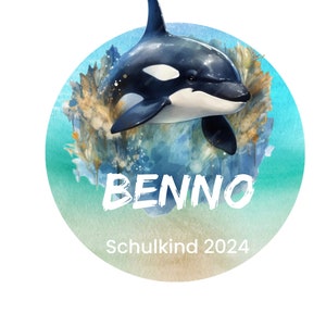 Può includere: Un'illustrazione ad acquerello di un'orca che nuota nell'oceano. L'orca è nera e bianca con una macchia bianca sull'occhio. Lo sfondo è un acquerello blu e verde con un acquerello marrone chiaro in basso. Il testo "BENNO Schulkind 2024" è scritto in bianco in basso all'immagine.
