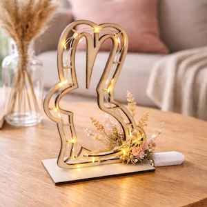 May include: A wooden bunny-shaped light with warm white LED lights. The bunny is on a rectangular base and decorated with dried flowers and small pink blossoms. The light is on a wooden table.