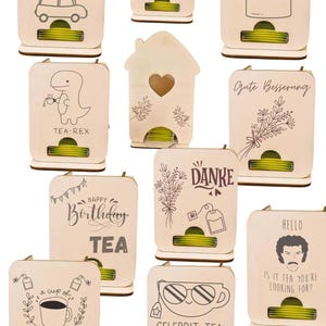 Peut inclure: Plusieurs distributeurs de sachets de thé en bois avec des motifs et des textes variés. Les motifs incluent une scène de Noël, un dinosaure avec une tasse à thé et les mots "TEA SHIRT". Chaque distributeur a une fente pour les sachets.