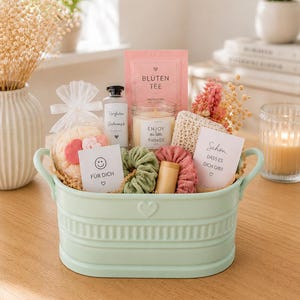 May include: A light green oval metal basket with handles containing a gift set. Items include a pink tea packet labeled BLÜTEN TEE, a candle labeled ENJOY the little THINGS, two fabric hair scrunchies, a bath sponge, and two cards with text.