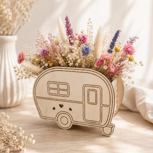 Könnte beinhalten: Eine holzgeschnitzte Wohnwagen-Vase, gefüllt mit einem bunten Arrangement aus Trockenblumen. Die Blumen umfassen rosa, lila, blaue und gelbe Blüten mit weißen und beigen Akzenten. Die Vase hat ein ausgeschnittenes Design eines Wohnwagens mit Tür, Fenstern und Herzdetails.