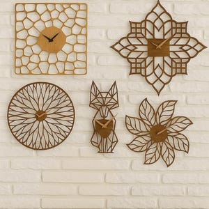 Puede incluir: Cinco relojes de pared de madera con diferentes diseños geométricos. Los relojes incluyen un cuadrado con un patrón de panal, una estrella, un círculo con líneas radiales, un zorro y una flor. Cada reloj tiene manecillas de madera y un acabado marrón claro.