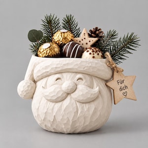 Puede incluir: Una maceta de cerámica color crema con forma de cabeza de Papá Noel, rellena de ramas de pino, una piña y chocolates. Una etiqueta de madera en forma de estrella atada con cuerda cuelga del lateral con el texto "Für dich" y un corazón.