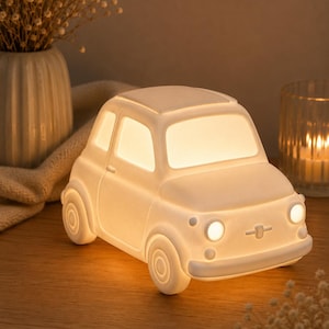 May include: A white, matte-finish car-shaped lamp that emits a warm, soft glow from its windows and circular headlights. The lamp features a compact, rounded design with a front bumper, side mirrors, and detailed wheel arches, resting on a flat surface.