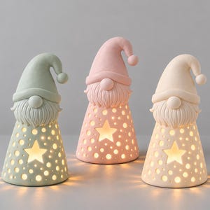 May include: Three conical gnome figurines in pastel sage green, soft pink, and cream. Each gnome features a pointed hat, a textured beard, and a base with cut-out star and circular patterns that emit a warm, internal glow from within the structure.