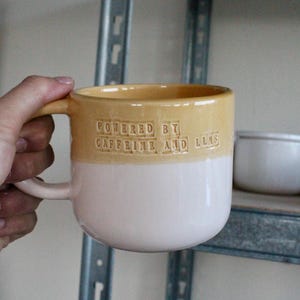 Può includere: Una tazza in ceramica bicolore con una sezione superiore giallo senape lucida e una sezione inferiore rosa chiaro liscia. Il testo "POWERED BY CAFFEINE AND LLAS" è impresso in rilievo sulla parte gialla della tazza. Presenta un manico abbinato.