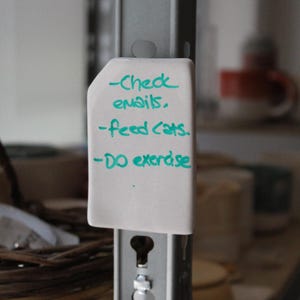 May include: A white, rectangular note is attached to a metal shelf support. The note has handwritten text in teal ink that reads: "-Check emails. -Feed cats. -DO exercise."