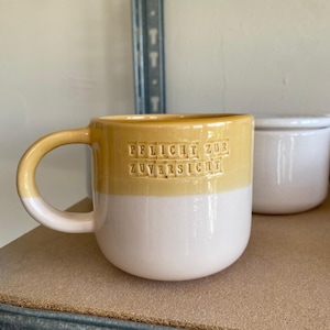 May include: A ceramic mug with a two-tone design featuring a mustard yellow upper section and a white lower section. The text "PFLICHT ZUR ZUVERSICHT" is embossed in the yellow portion. The mug has a curved handle and a rounded body shape.