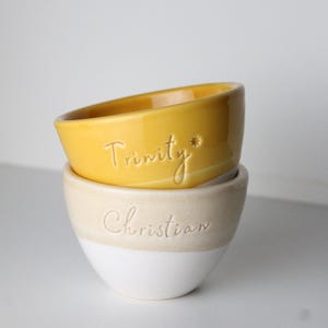 Puede incluir: Dos cuencos de cerámica apilados. El cuenco superior es amarillo con la palabra "Trinity" inscrita. El cuenco inferior es blanco con una banda beige y la palabra "Christian" inscrita. Los cuencos tienen un diseño sencillo. Medidas no disponibles.
