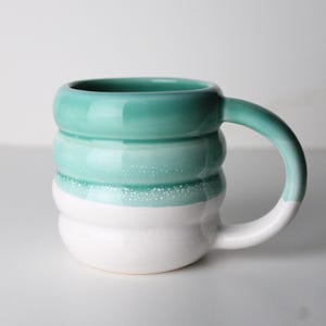 May include: A handcrafted ceramic mug with a distinctive, ridged pattern. The mug showcases a colour gradient, shifting from teal green at the top to white at the base. The handle is also teal green and white. Ideal for tea or coffee.