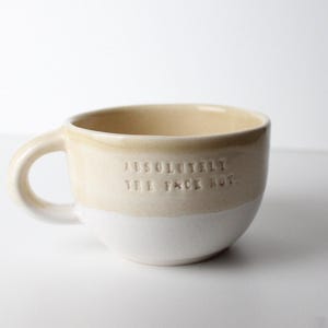 Pode incluir: Uma caneca de cerâmica com acabamento bicolor, apresentando uma seção superior bege e uma seção inferior branca. O texto "ABSOLUTELY THE F*CK NOT" está gravado em letras maiúsculas na parte frontal da caneca, que possui uma alça.