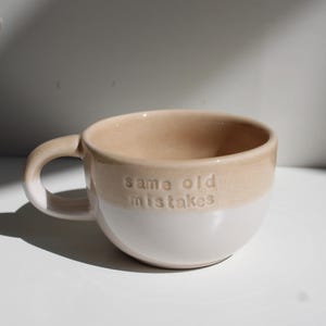 Könnte beinhalten: Ein zweifarbiger Keramikbecher mit einem beigen oberen Teil und einem weißen unteren Teil. Der Text "same old mistakes" ist in den beigen Bereich des Bechers eingeprägt. Er verfügt über einen geschwungenen Henkel an der linken Seite und einen Rand.