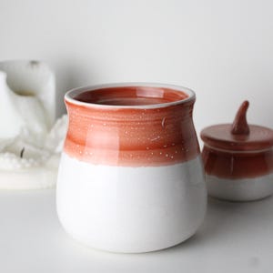Puede incluir: Un tarro de cerámica blanco con una abertura ancha y una base redondeada, acentuado con una banda de pintura naranja-roja. Un tarro más pequeño con tapa a juego está al lado. Los tarros tienen un acabado moteado.