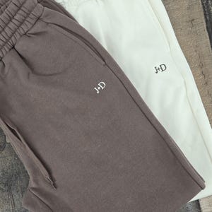 May include: Two pairs of sweatpants, one brown and one white, with the letters "J+D" embroidered on the front of each.